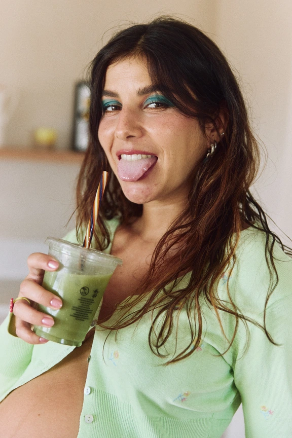 Mujer embarazada sonriendo mientras sostiene un vaso con una bebida verde.